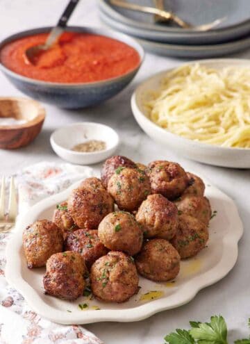 A platter of air fryer turkey meatballs with sauce and noodles in the back along with salt and pepper and plates.