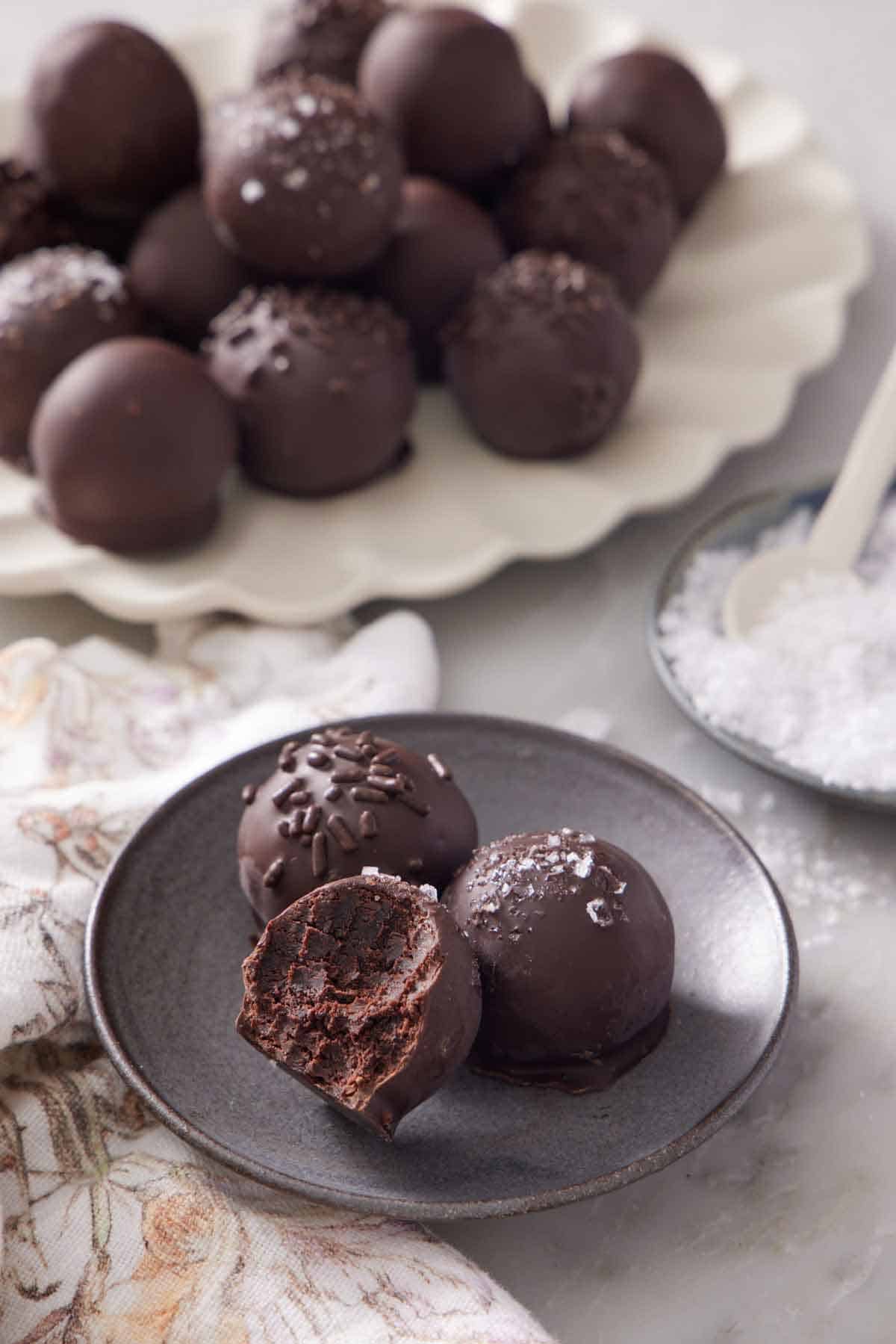 A plate with two and a half brownie truffles. A platter of more brownie truffles in the background along with a bowl of flaky salt.