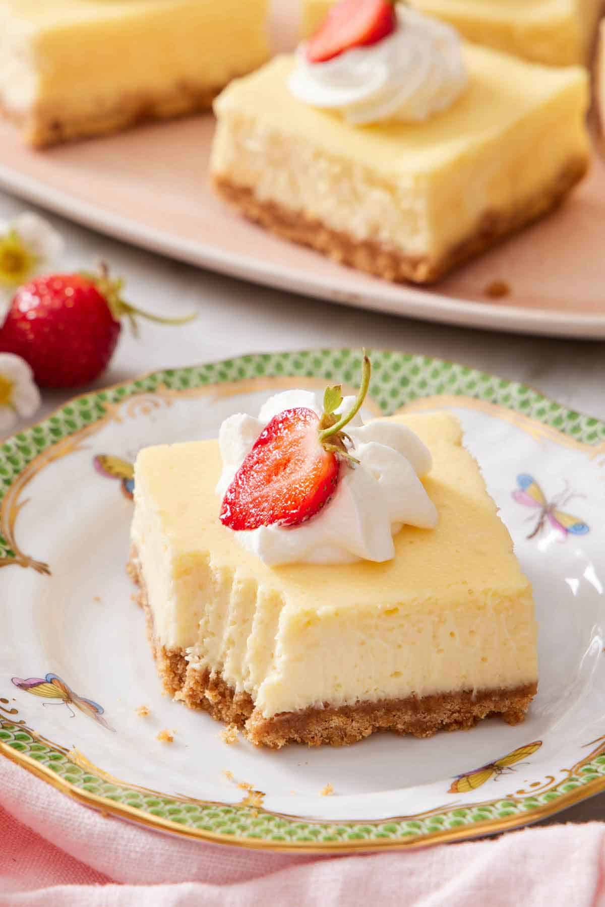 A plate with a cheesecake bar topped with whipped cream and strawberry with a bite taken out of the bar.