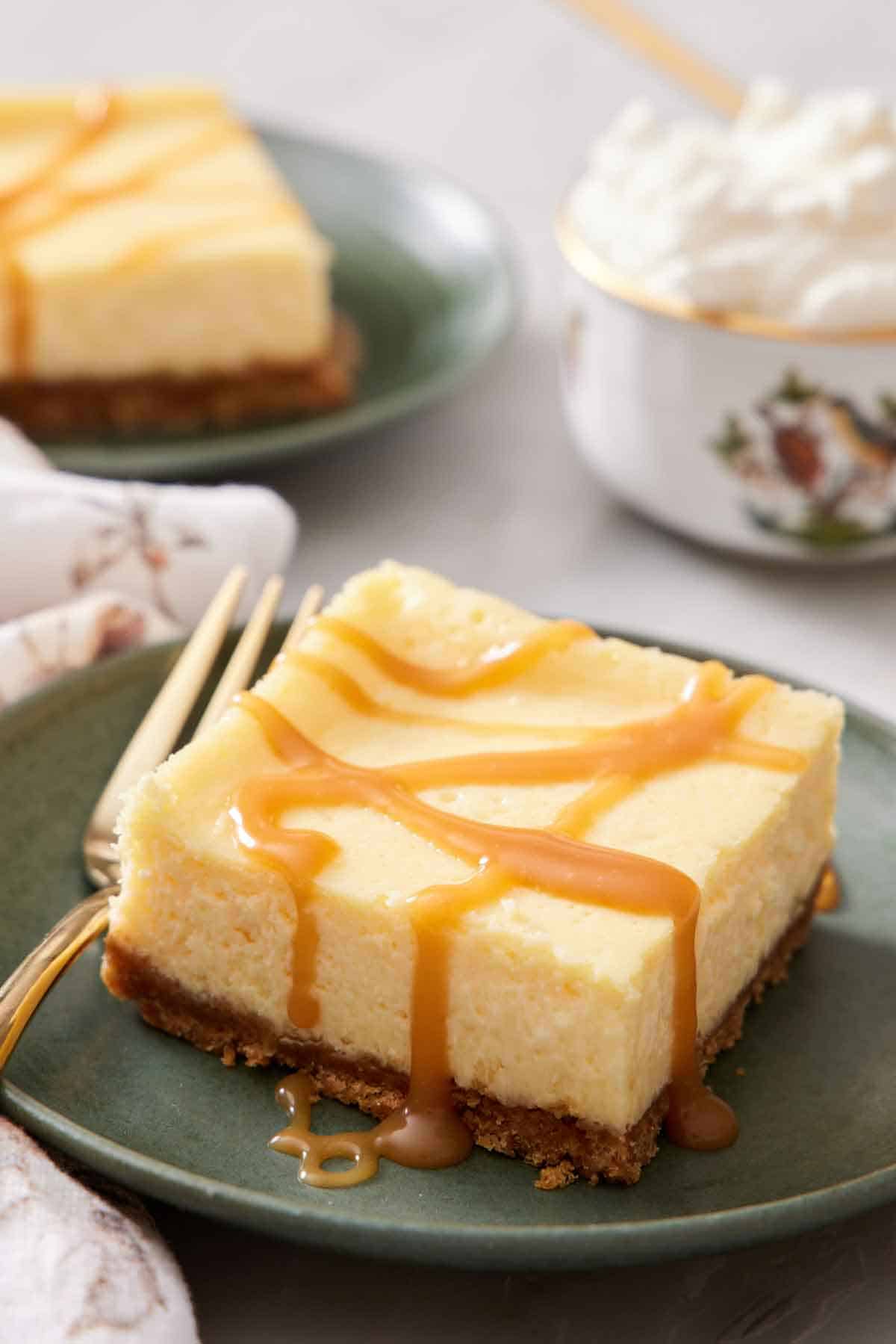 A plate with a cheesecake bar topped with a drizzle of caramel. Whipped cream in the background and another plate with another bar.