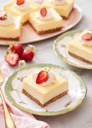 A plate with a cheesecake bar with whipped cream and strawberry on top. More cheesecake bars in the background with strawberries.