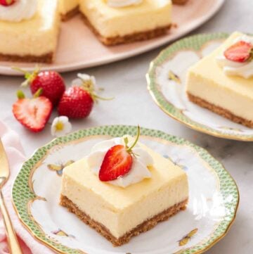 A plate with a cheesecake bar with whipped cream and strawberry on top. More cheesecake bars in the background with strawberries.