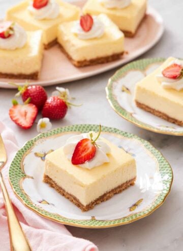 A plate with a cheesecake bar with whipped cream and strawberry on top. More cheesecake bars in the background with strawberries.