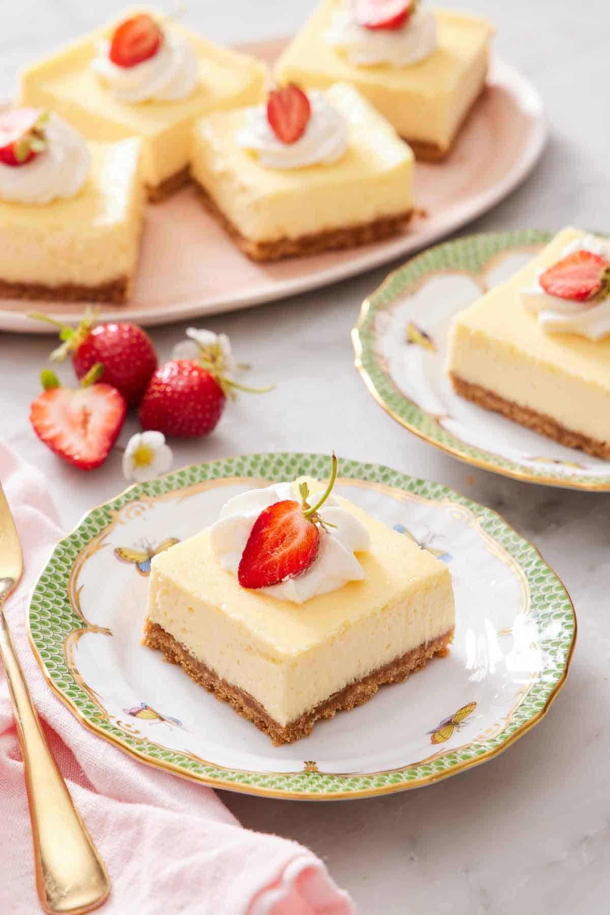 A plate with a cheesecake bar with whipped cream and strawberry on top. More cheesecake bars in the background with strawberries.