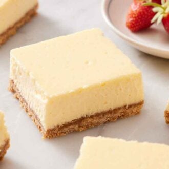 Cheesecake bars scattered on a marble surface. A small plate of strawberries in the background.