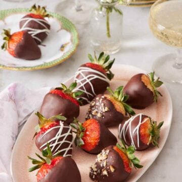 A platter of chocolate covered strawberries with a small vase of flowers and small plate with two chocolate covered strawberries in the background.