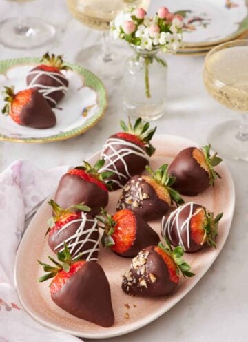 A platter of chocolate covered strawberries with a small vase of flowers and small plate with two chocolate covered strawberries in the background.