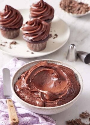 A bowl of chocolate cream cheese frosting with three frosted cupcakes in the background. Piping tips on the side.