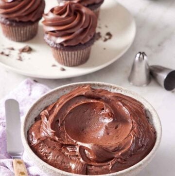 A bowl of chocolate cream cheese frosting with three frosted cupcakes in the background. Piping tips on the side.