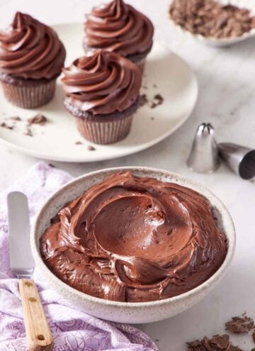 A bowl of chocolate cream cheese frosting with three frosted cupcakes in the background. Piping tips on the side.