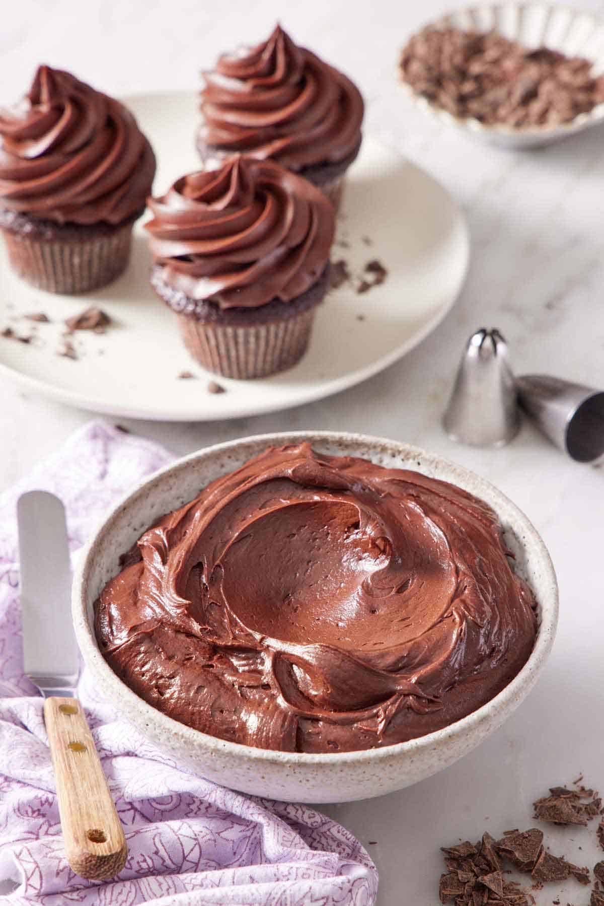 A bowl of chocolate cream cheese frosting with three frosted cupcakes in the background. Piping tips on the side.