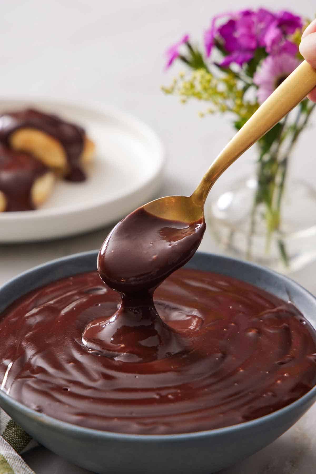 A spoon lifting up a spoonful of chocolate gravy from a bowl. A vase of flowers in the background.