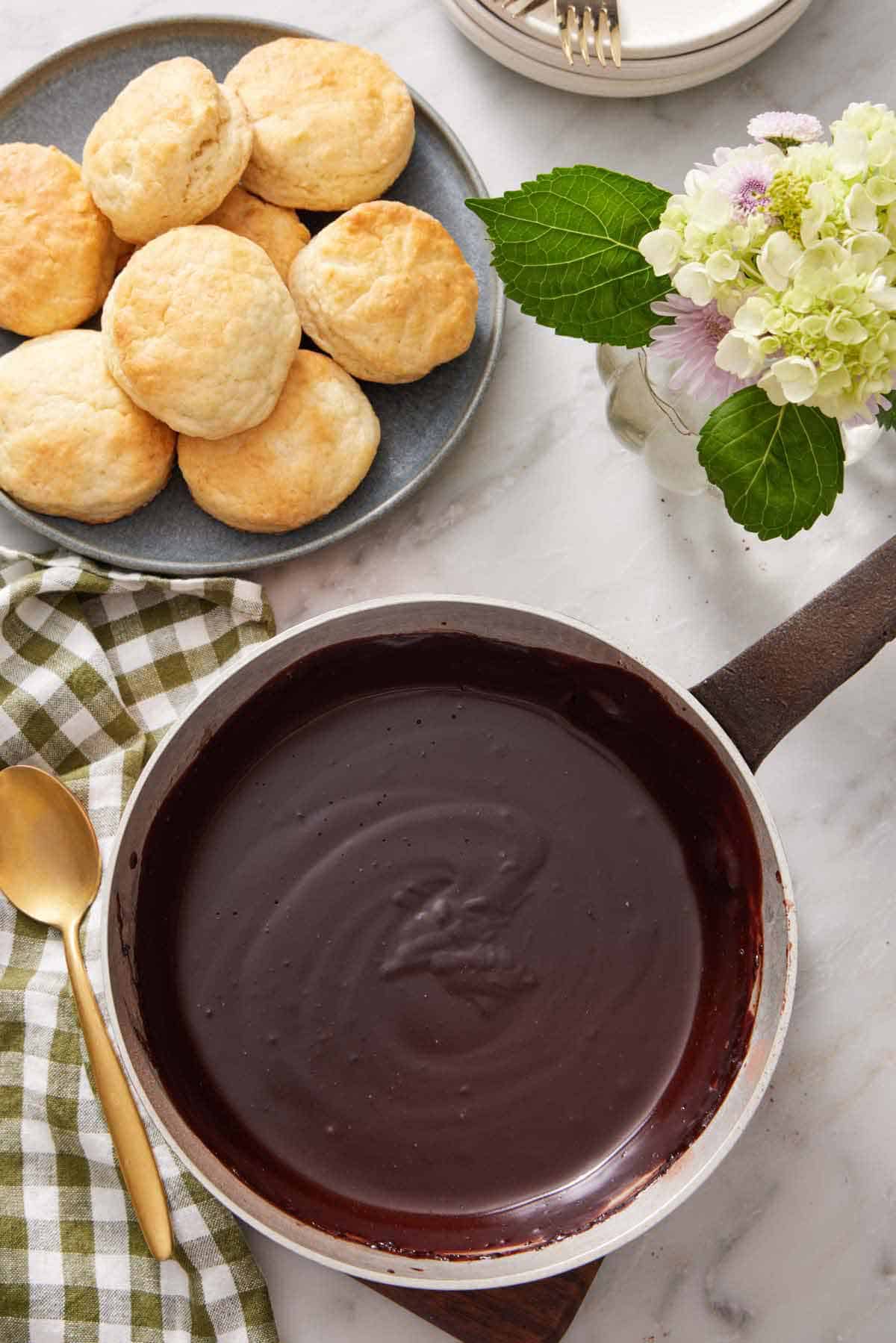 Overhead view of a pot of chocolate gravy. A plate of biscuits, spoon, and vase of flower beside it.