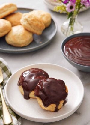 A plate with two biscuits with chocolate gravy on top. A bowl of chocolate gravy in the back along with a plate of biscuits and vase of flowers.