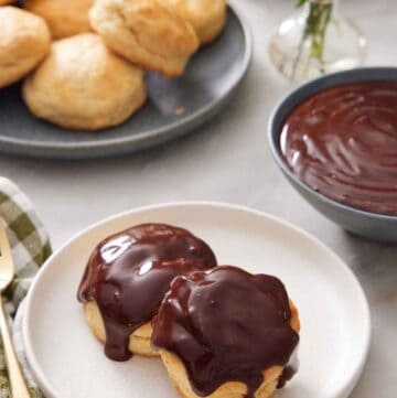 A plate with two biscuits with chocolate gravy on top. A bowl of chocolate gravy in the back along with a plate of biscuits and vase of flowers.