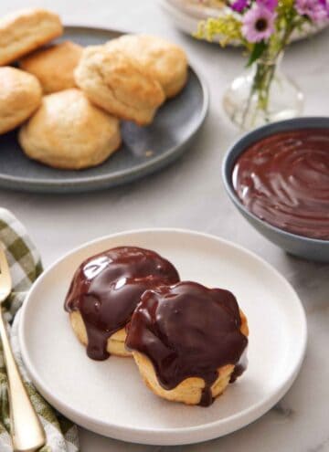 A plate with two biscuits with chocolate gravy on top. A bowl of chocolate gravy in the back along with a plate of biscuits and vase of flowers.