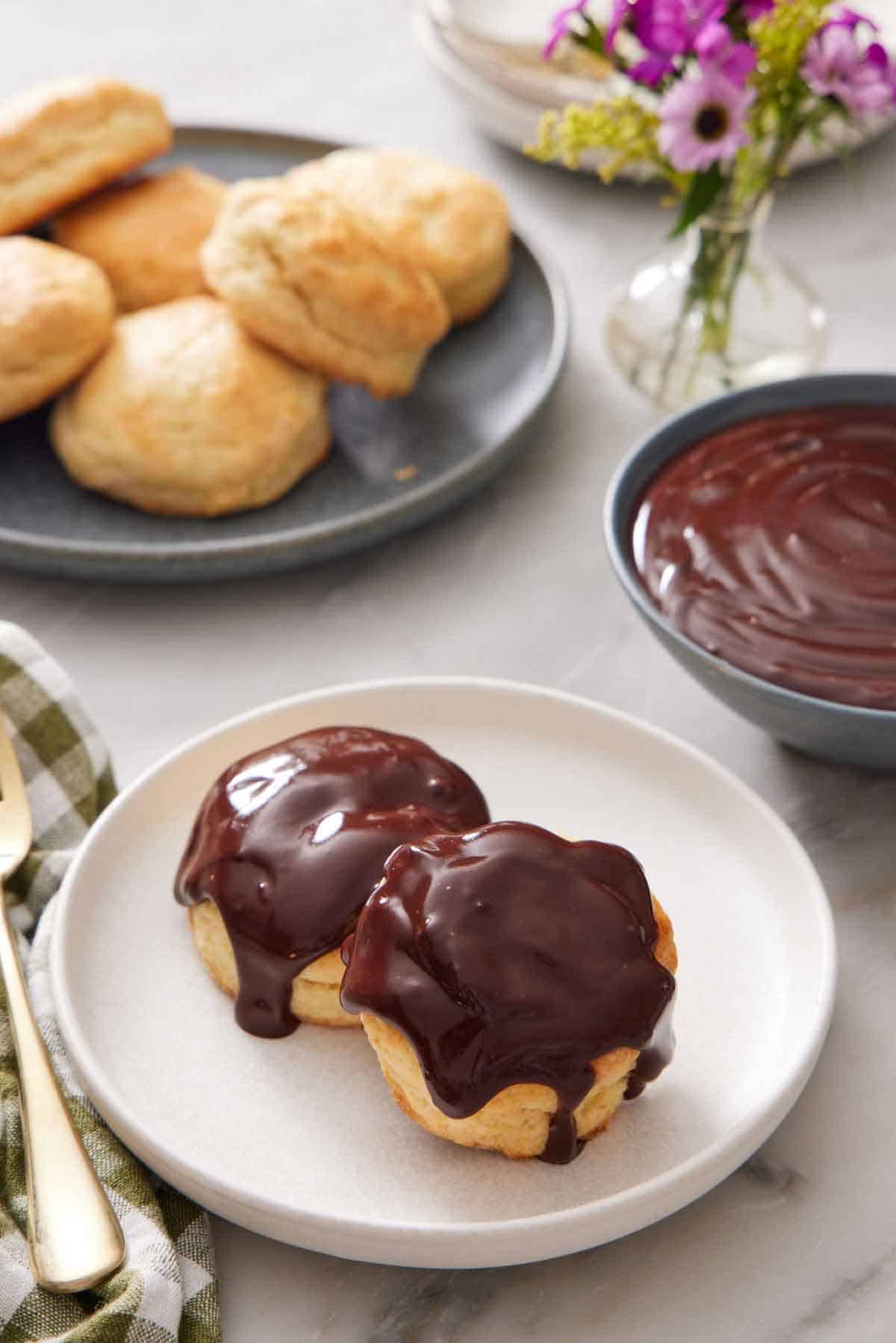 A plate with two biscuits with chocolate gravy on top. A bowl of chocolate gravy in the back along with a plate of biscuits and vase of flowers.