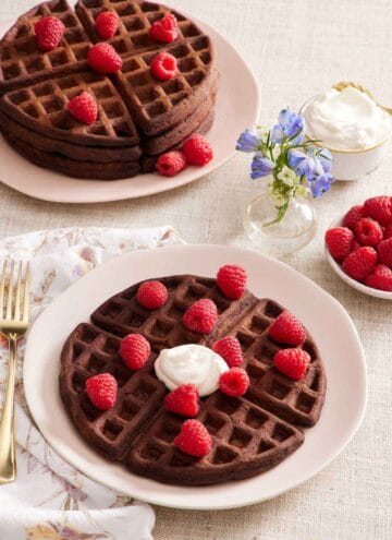 A plate with a chocolate waffle topped with some whipped cream and fresh raspberries. A stack of more waffles topped with raspberries, a bowl of raspberries, a vase of flowers, and a bowl of whipped cream in the back.