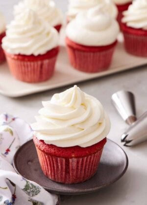 A red velvet cupcake with ermine frosting. More frosted cupcakes in the background and piping tips.