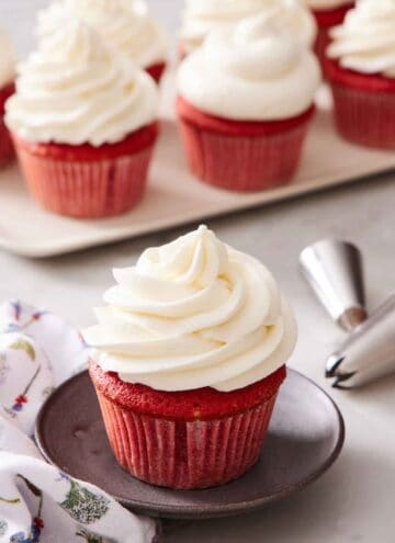 A red velvet cupcake with ermine frosting. More frosted cupcakes in the background and piping tips.