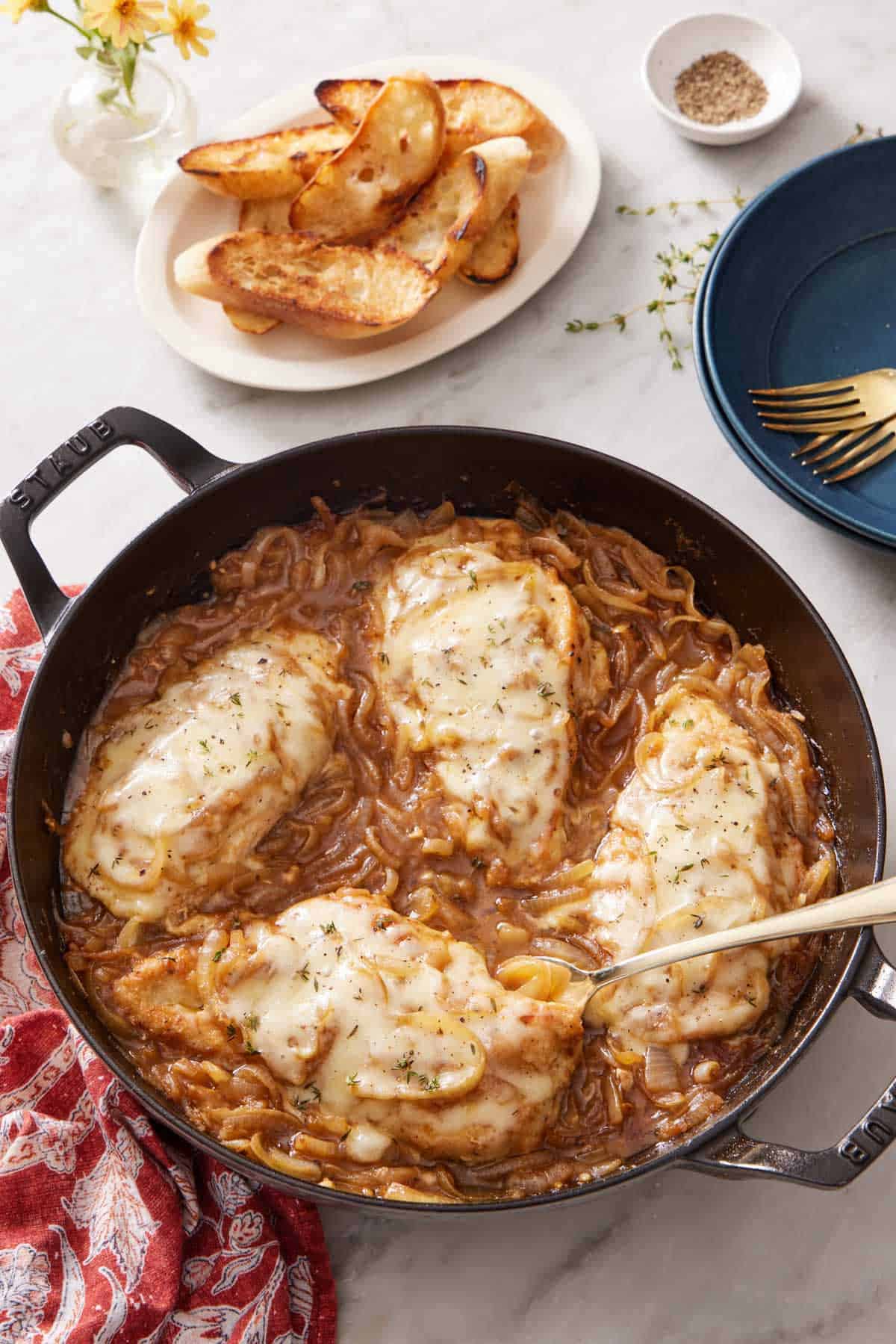 Overhead view of a skillet of French onion chicken with a spoon tucked in underneath one chicken breast. A small plate of toasted bread in the background with some pepper, forks, and plates.