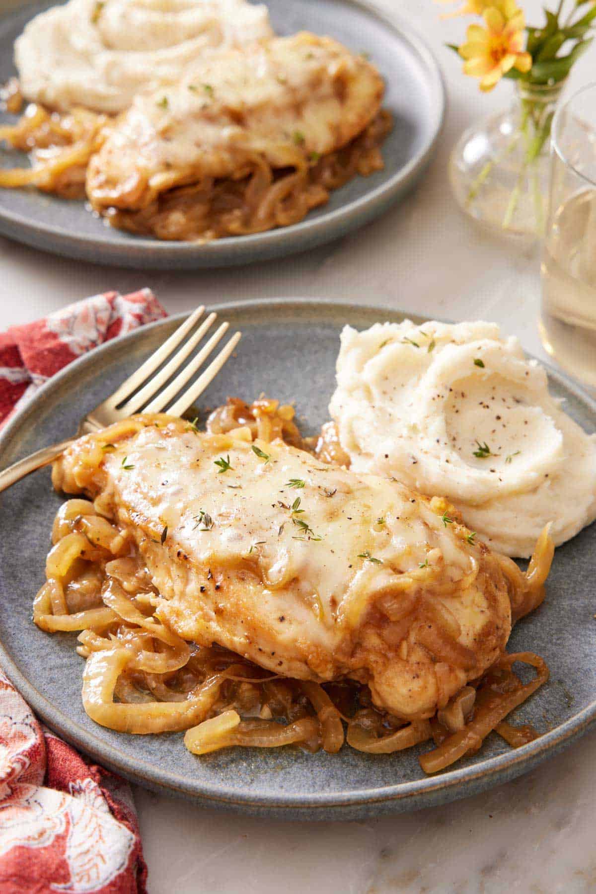 A plate of French onion chicken with a fork and mashed potatoes. Another plated serving in the background.