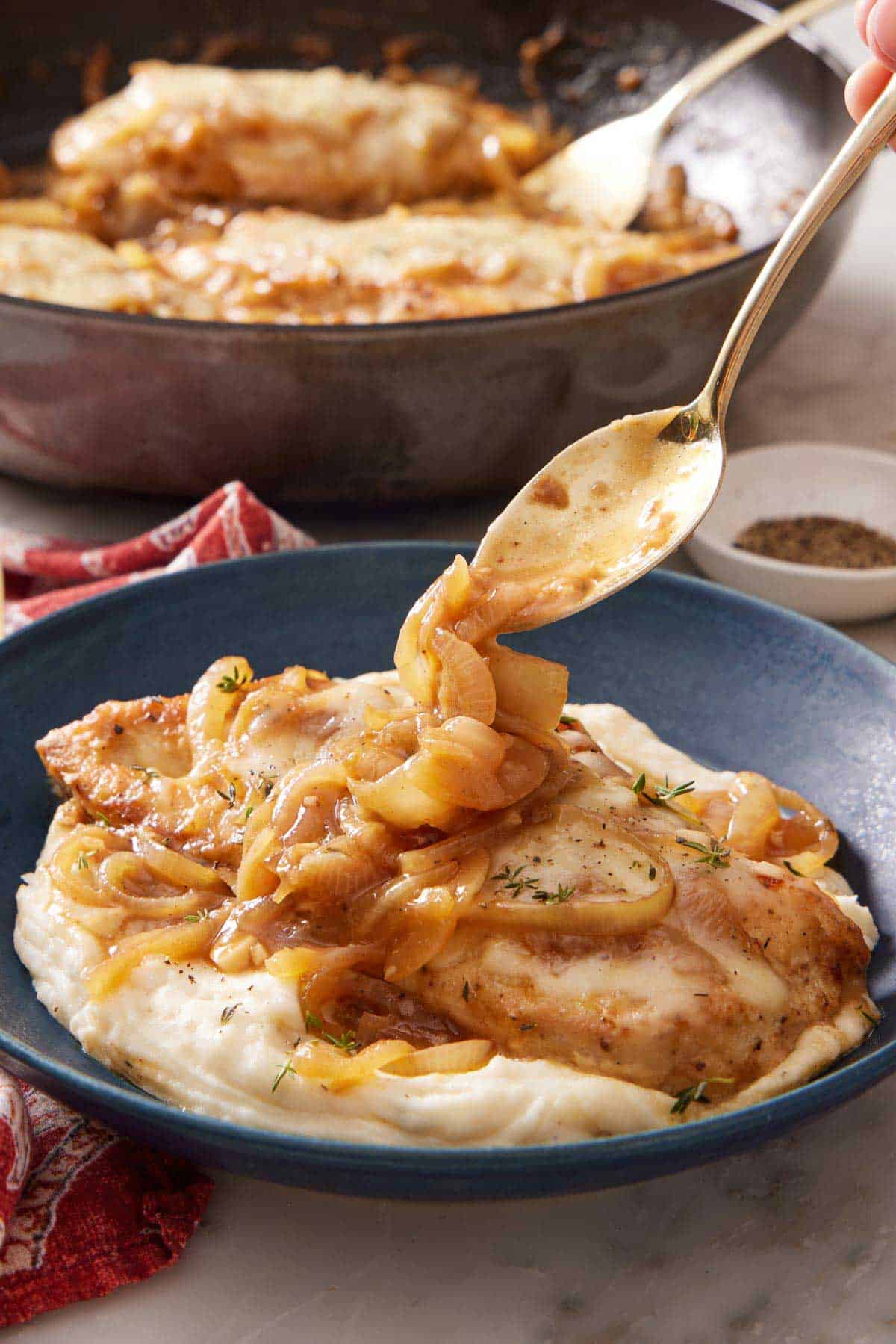 A spoon spooning some onions over French onion chicken over mashed potatoes in a bowl. A skillet of more chicken in the background.