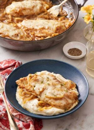 A plate of French onion chicken over mashed potatoes. A skillet with more chicken in the background.