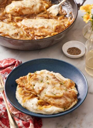 A plate of French onion chicken over mashed potatoes. A skillet with more chicken in the background.