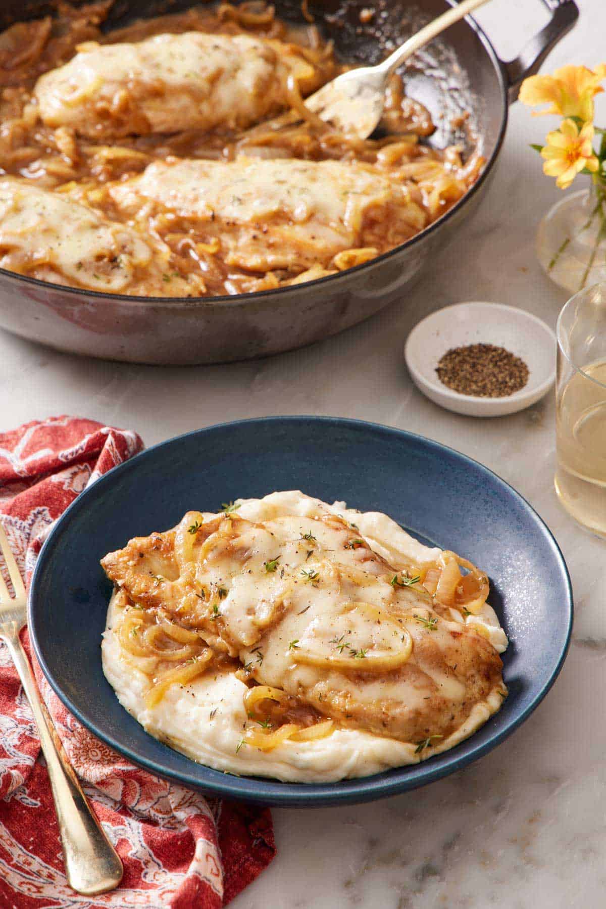 A plate of French onion chicken over mashed potatoes. A skillet with more chicken in the background.