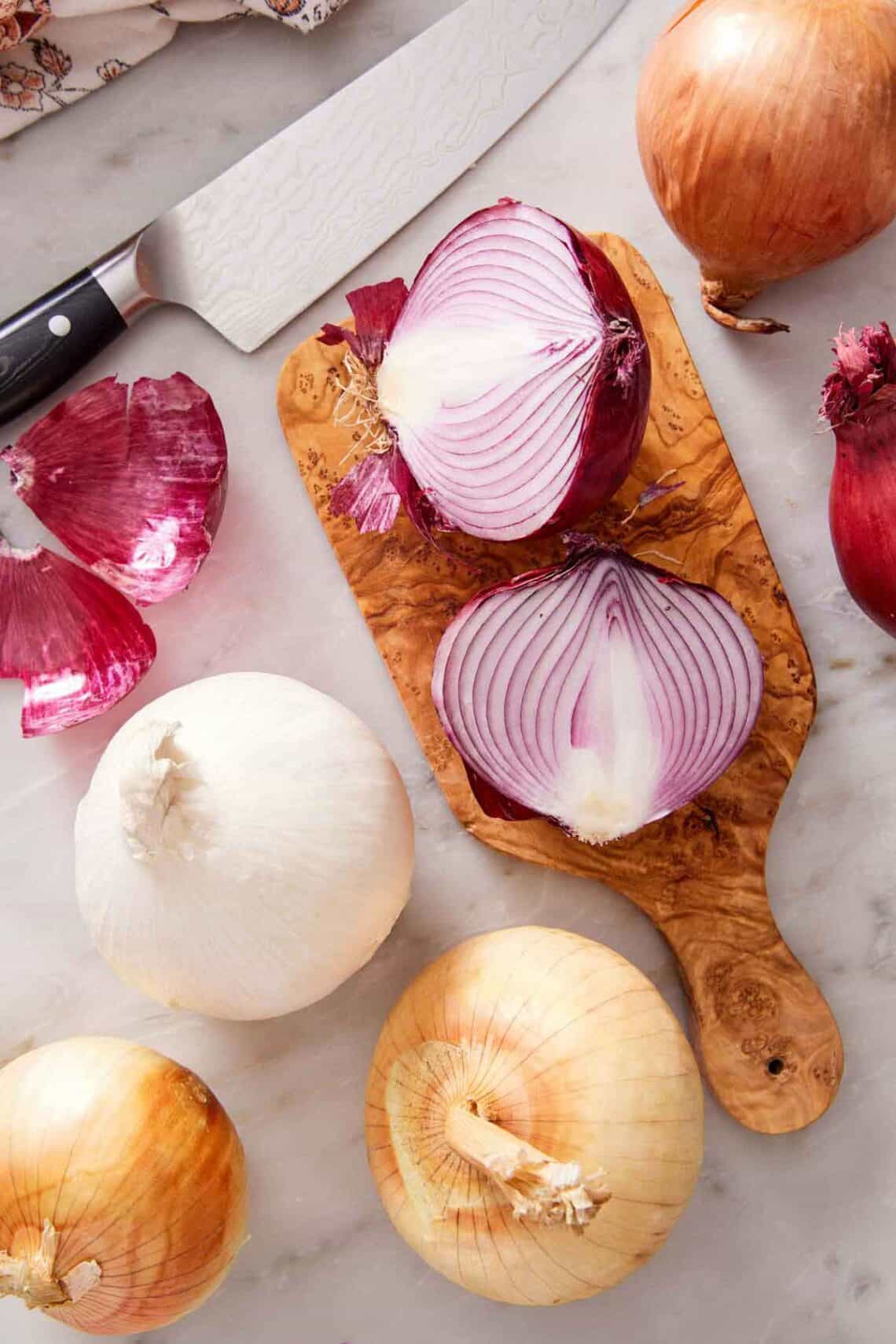 Overhead view of a cutting board with a red onion cut in half and yellow and white onions scattered around.