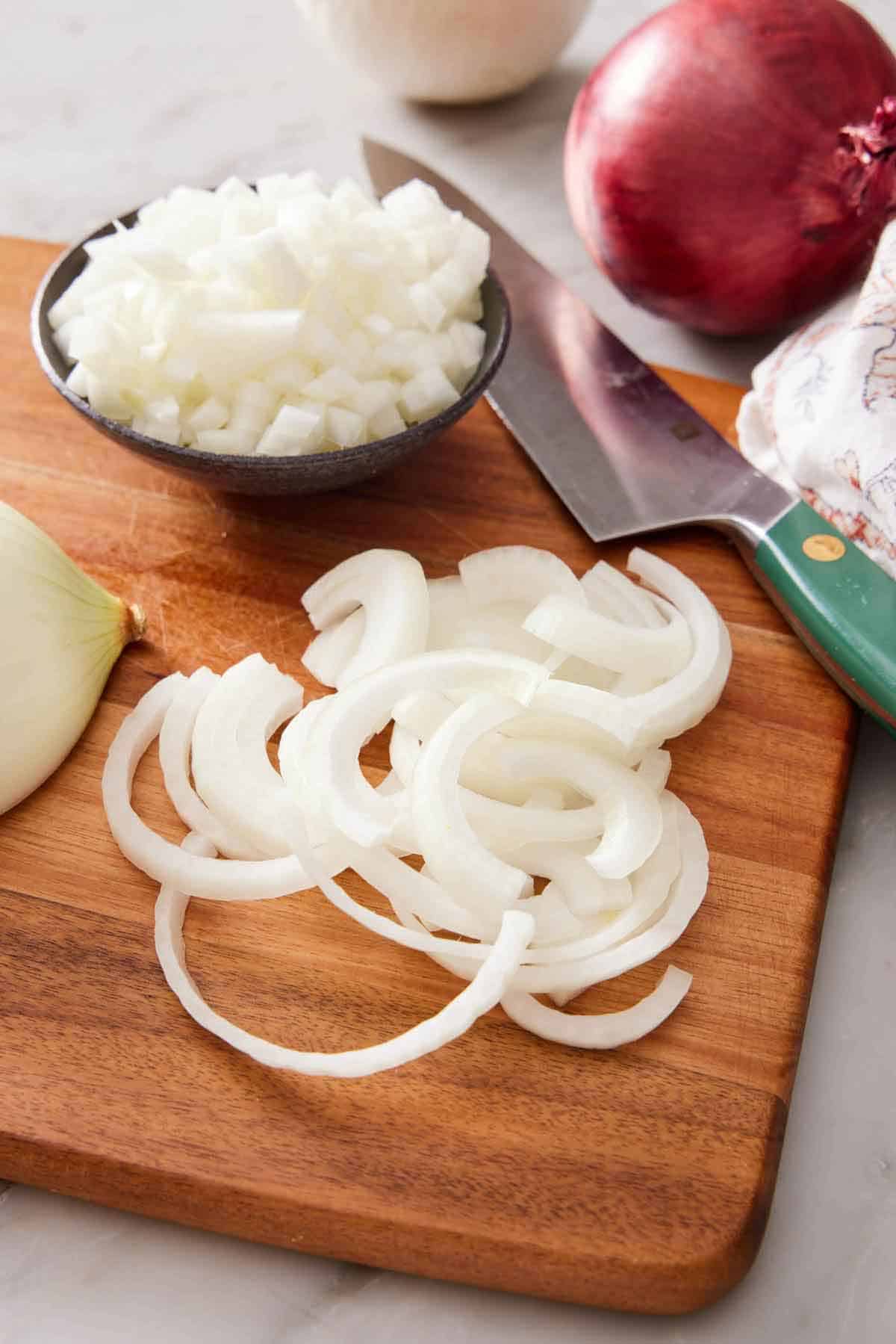 A cutting board with sliced onions, a knife, and a bowl of diced onions. A red onion in the background.