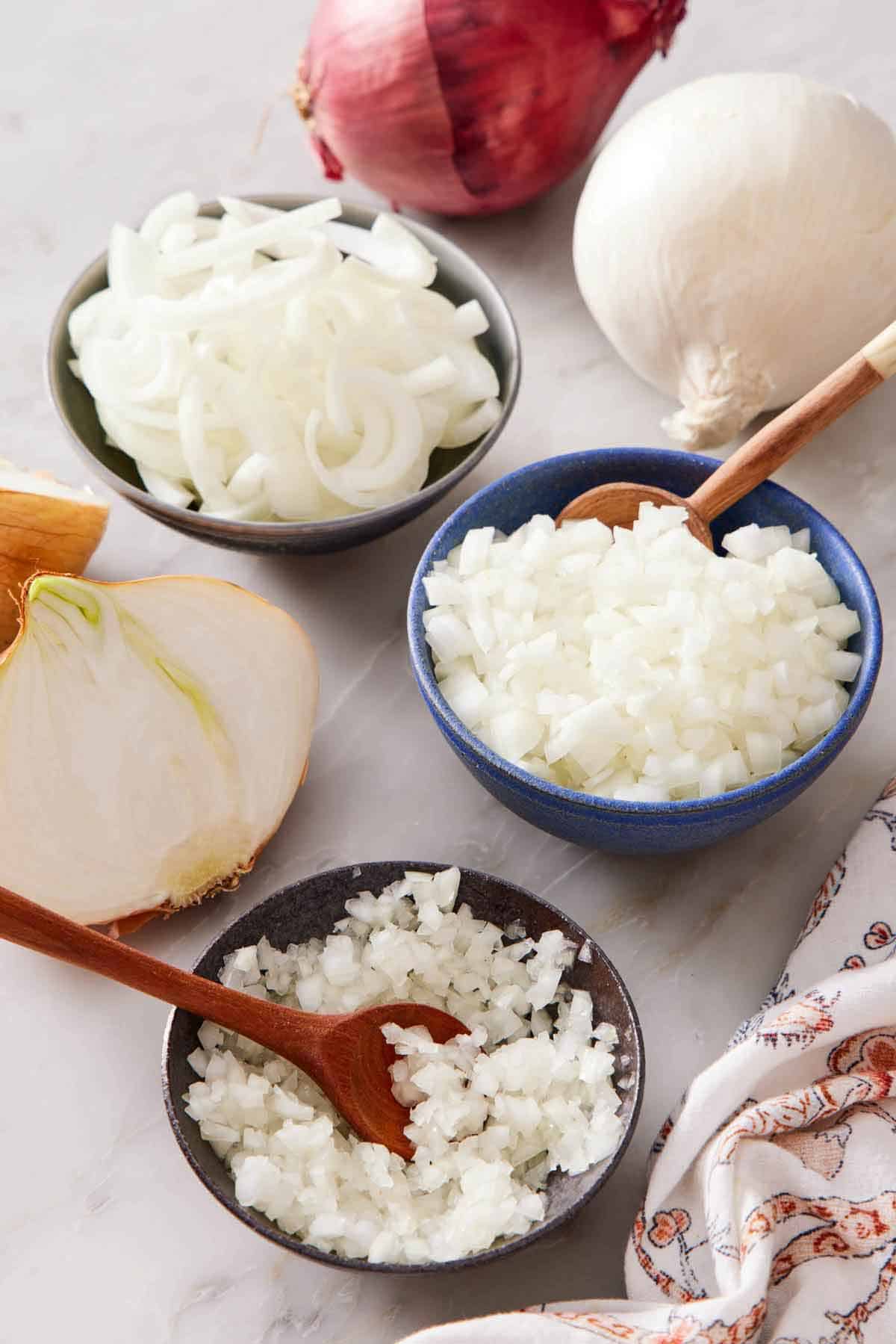 A bowl of sliced onions, diced onions, and finely diced onions. A red onion, white onion, and cut yellow onion scattered around.