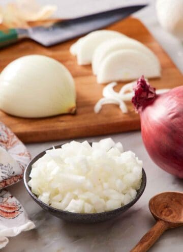 A bowl of diced onions with another onion in the background on a cutting board in the process of being cut and a red onion on the side.