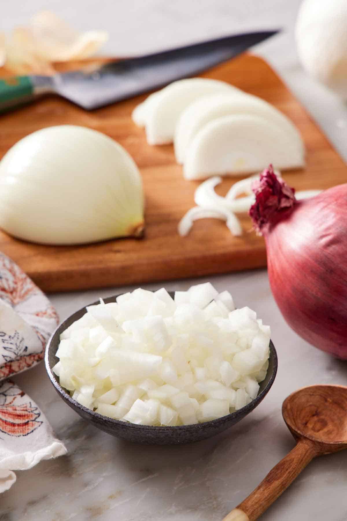 A bowl of diced onions with another onion in the background on a cutting board in the process of being cut and a red onion on the side.