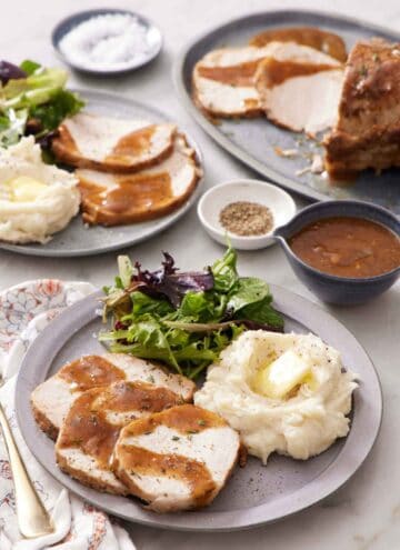 A plate with a serving of Instant Pot pork loin with gravy, mashed potatoes, and salad. Another plated serving in the back along with the rest of the pork, a bowl of gravy, salt, and pepper.