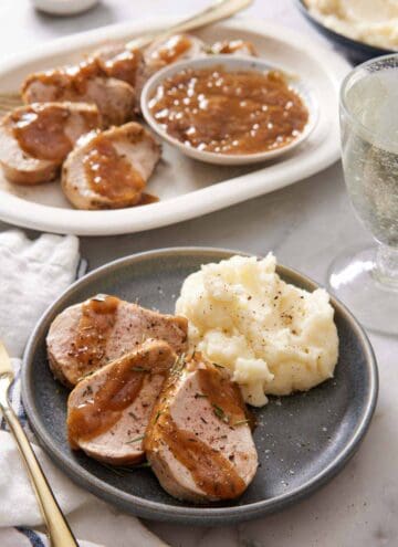 A plate with three pieces of Instant Pot pork tenderloin on a plate with mashed potatoes. A platter with more sliced tenderloin in the background along with a bowl of gravy.