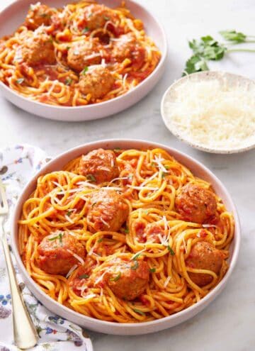 A plate of Instant Pot spaghetti and meatballs topped with parsley and parmesan. A bowl of parmesan and another plated serving of spaghetti and meatballs in the background.