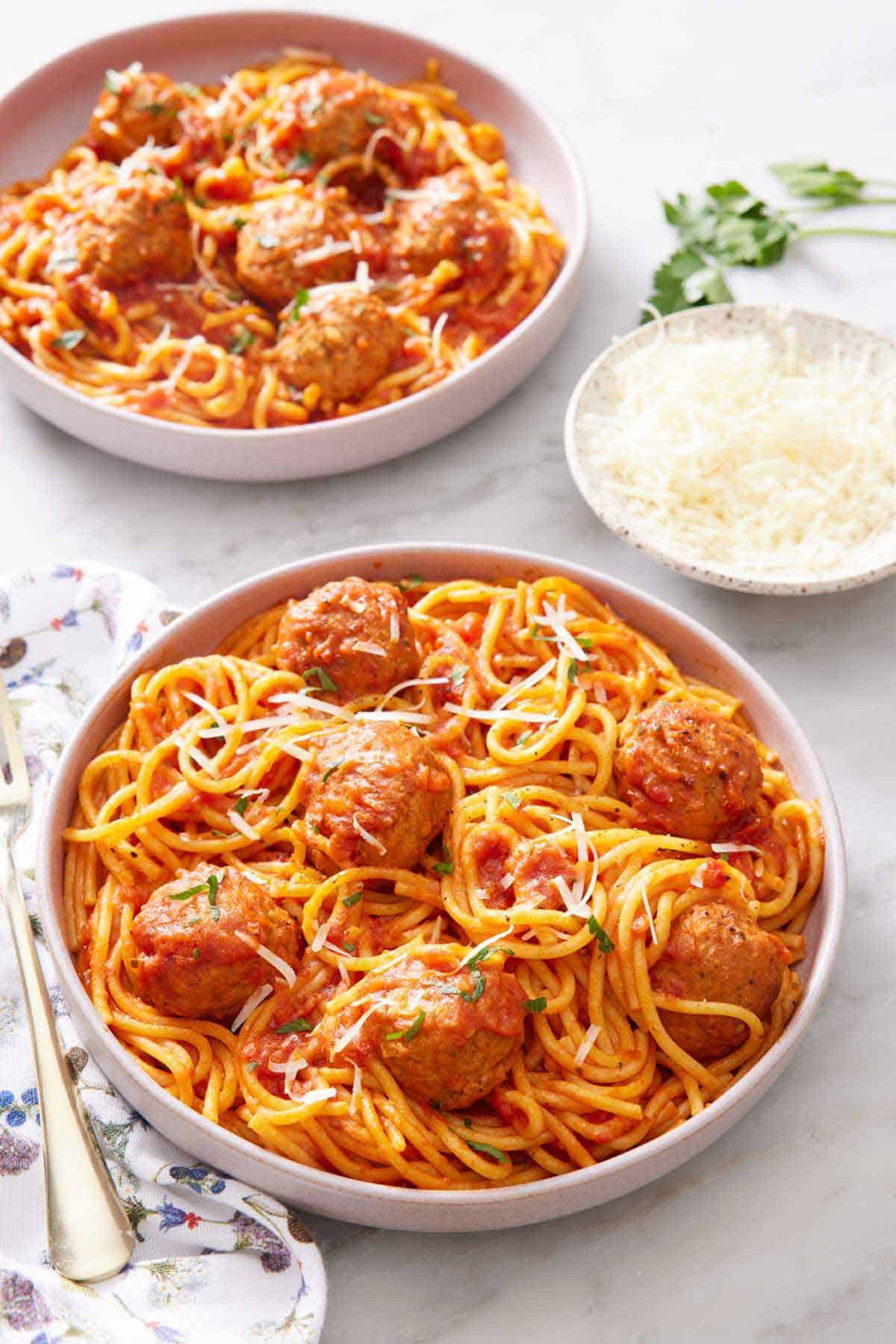 A plate of Instant Pot spaghetti and meatballs topped with parsley and parmesan. A bowl of parmesan and another plated serving of spaghetti and meatballs in the background.