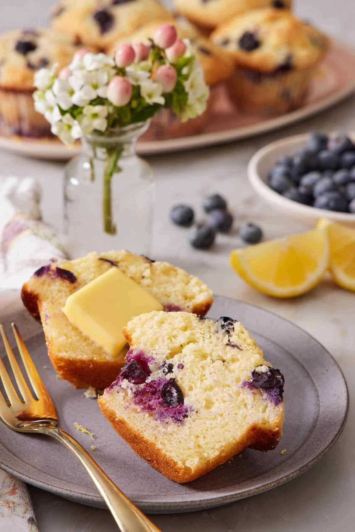 A plate with a fork with a lemon blueberry muffin cut in half with a slab of butter on one half. A mini vase of flowers, lemon wedges, blueberries, and more muffins in the background.