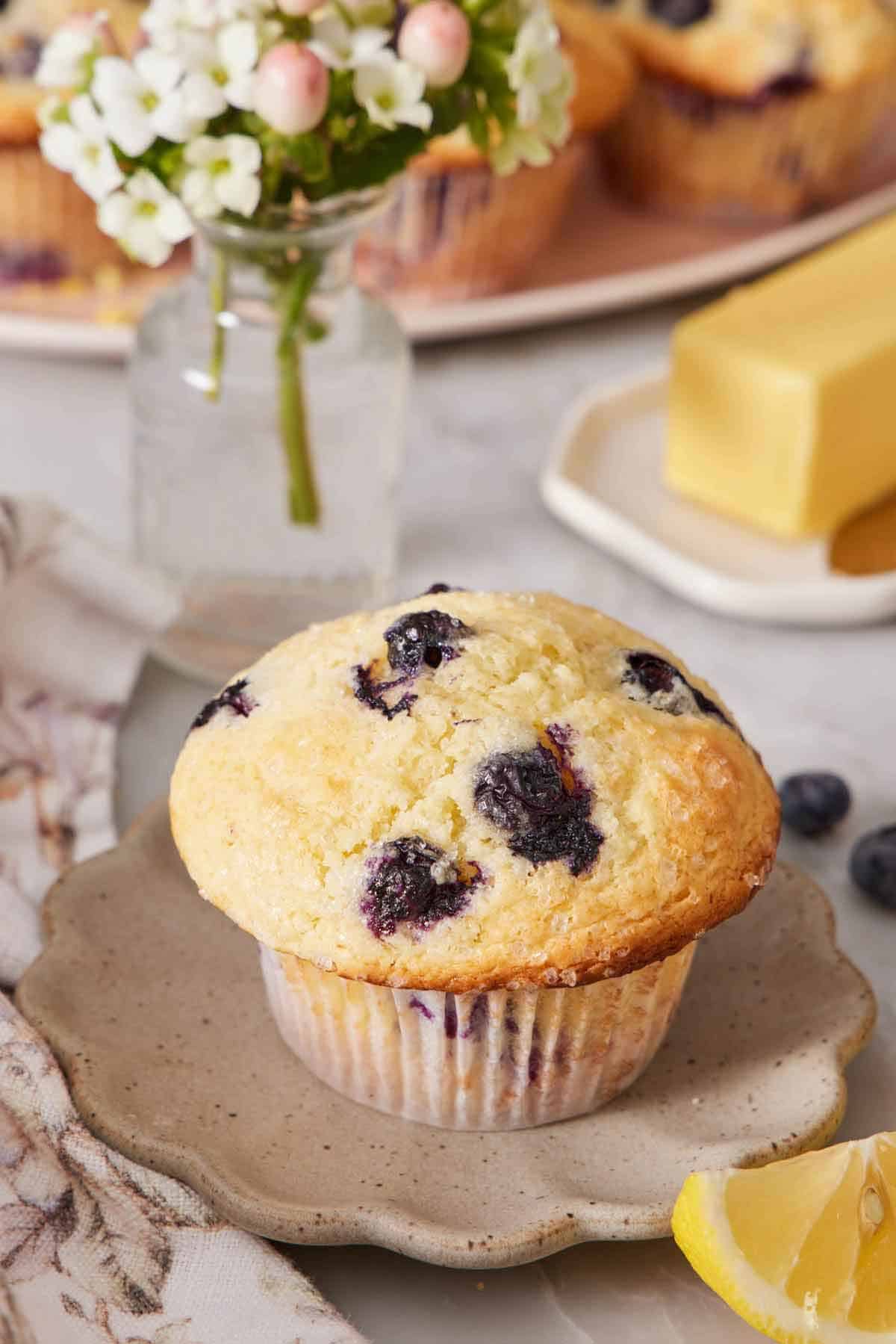 A small scalloped plate with a lemon blueberry muffin. A mini vase of flowers, lemon wedges, blueberries, and butter scattered around.