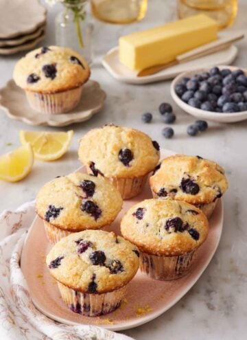 A platter of lemon blueberry muffins. A bowl of blueberries, butter, lemon wedges, and a mini plate with another muffin on it in the background.