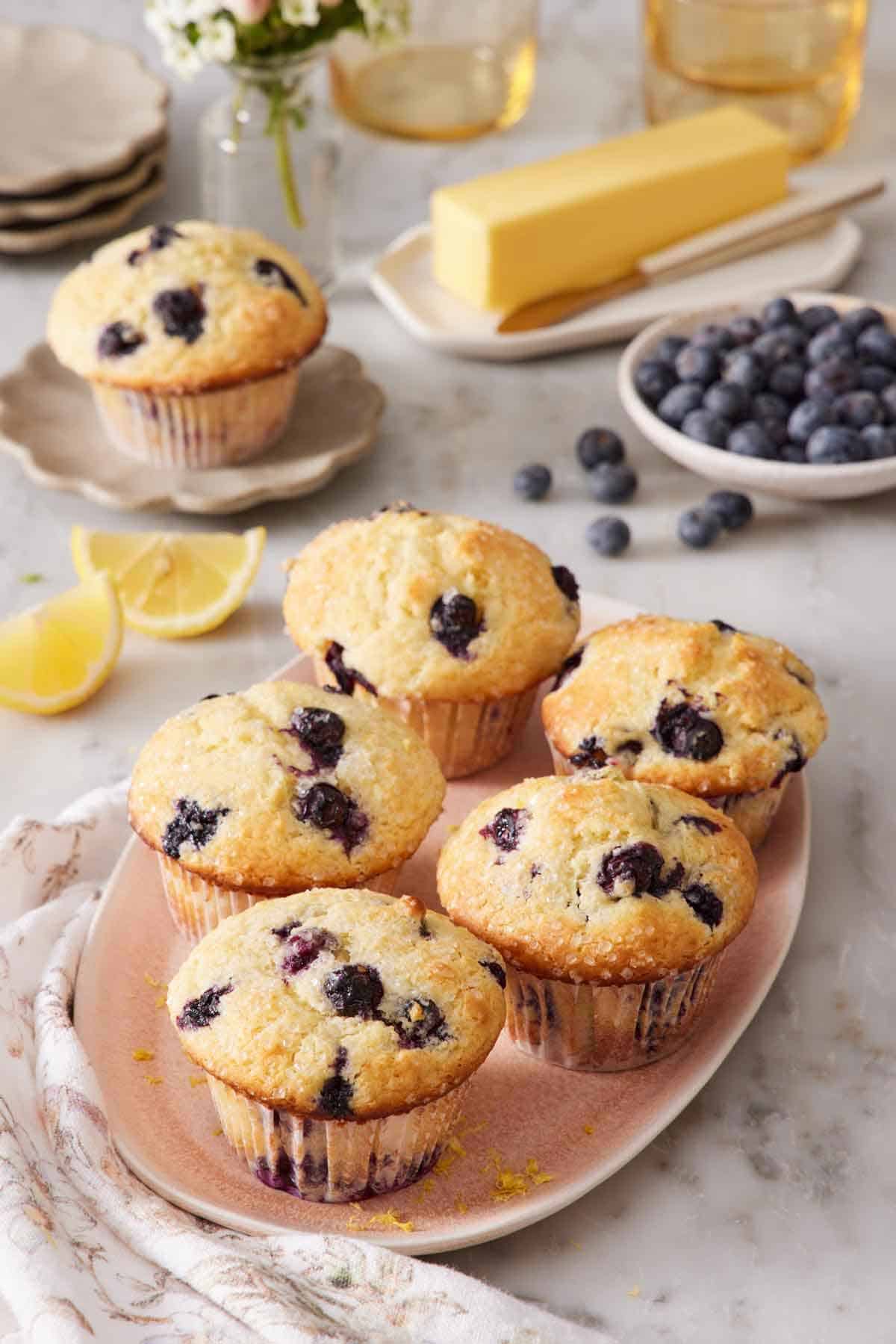 A platter of lemon blueberry muffins. A bowl of blueberries, butter, lemon wedges, and a mini plate with another muffin on it in the background.