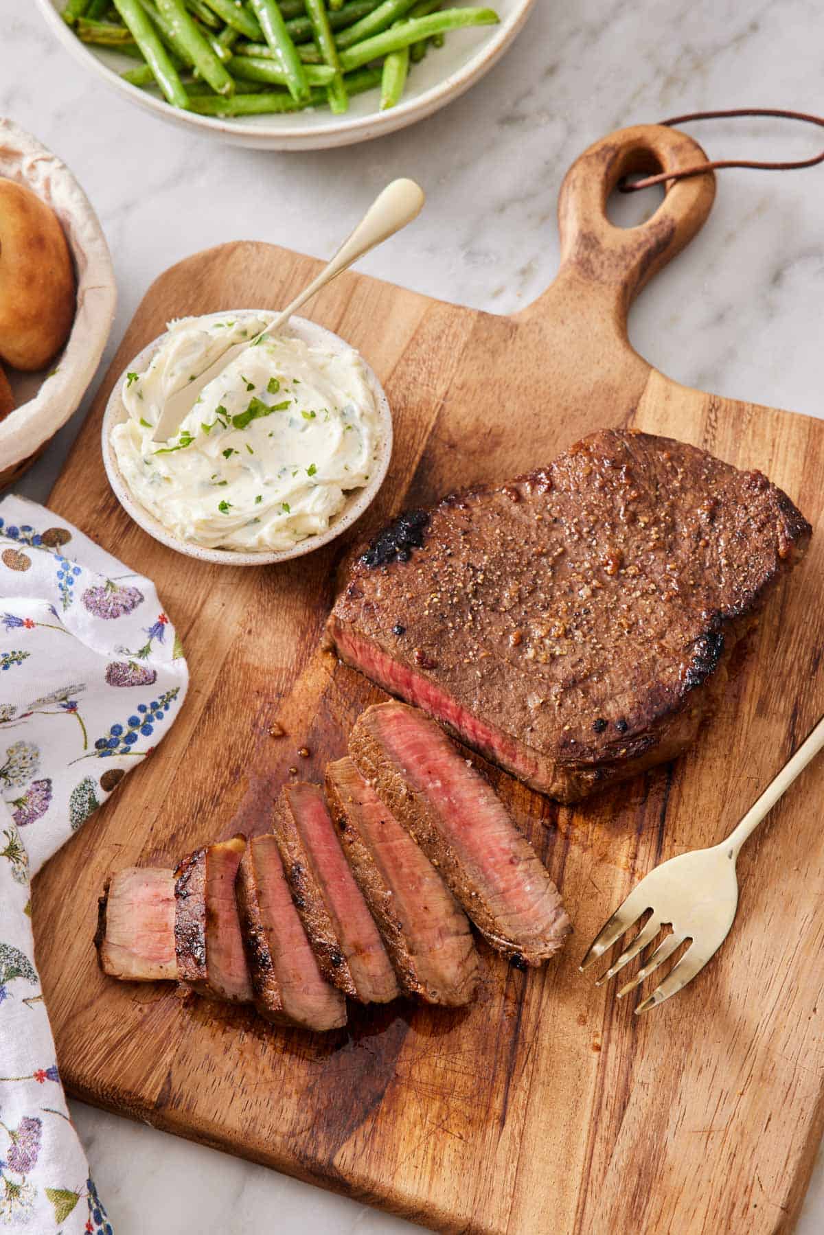 A wooden cutting board with a London broil, half sliced. A fork and a bowl of herb compound butter beside the steak.