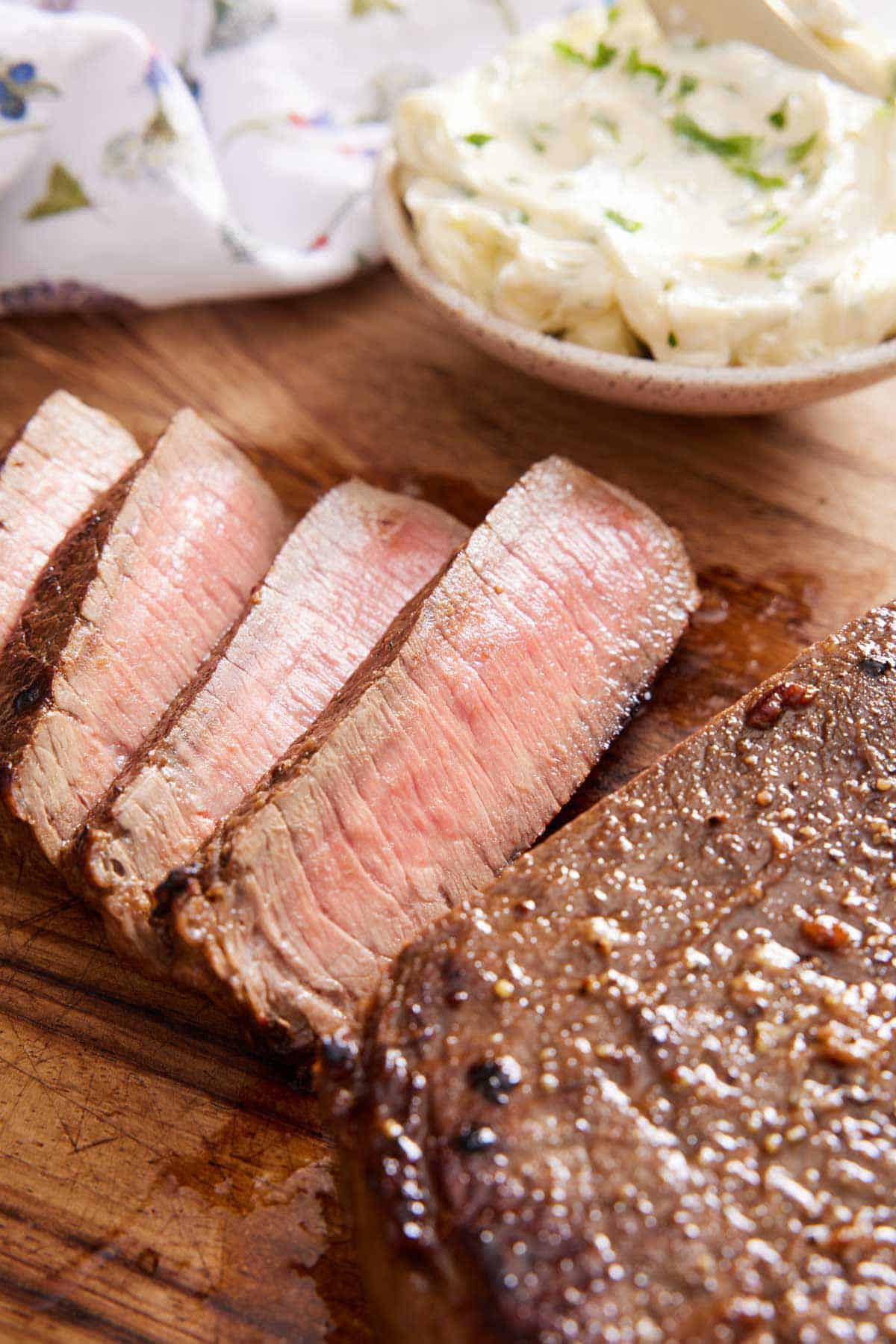 A close up view of sliced London broil on a wooden cutting board. A bowl of herb butter int he back.