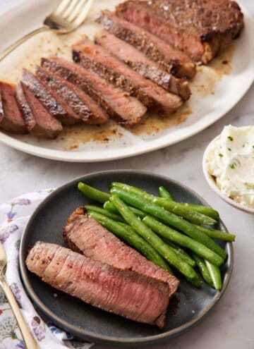 A plate with two slices of London broil with green beans. A platter in the background with the rest of the sliced London broil.