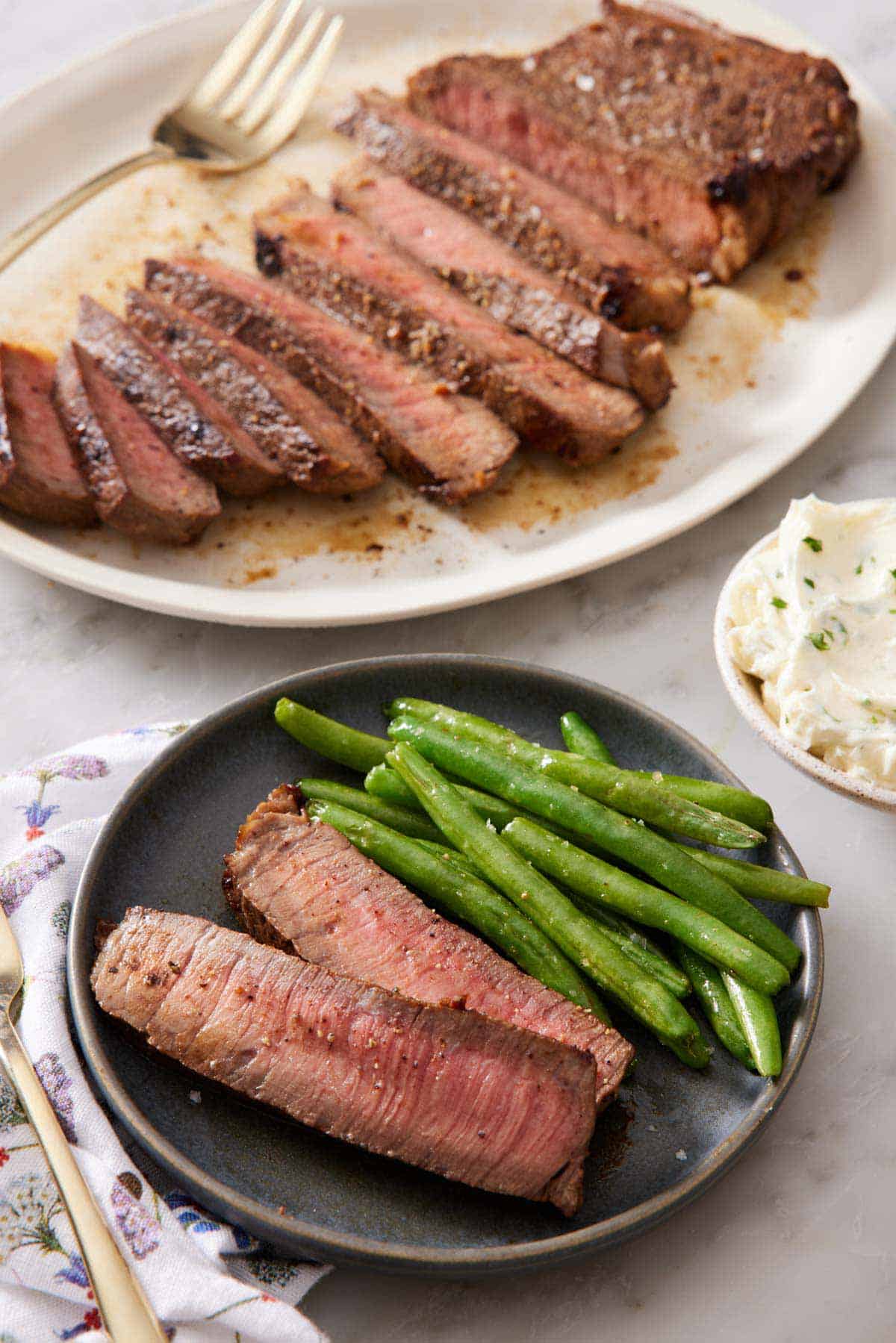 A plate with two slices of London broil with green beans. A platter in the background with the rest of the sliced London broil.