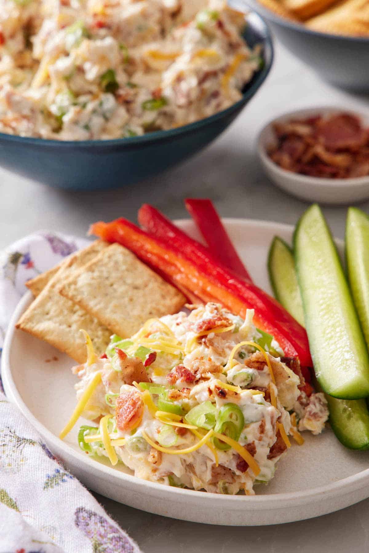 A plate with a serving of million dollar dip along with crackers, pepper peppers, and cucumbers. A bowl of dip in the background.