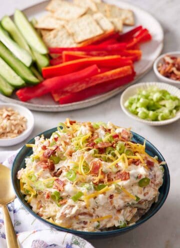 A bowl of million dollar dip with a platter in the background with cucumbers, peppers, and crackers. More ingredients in individual bowls off to the side.