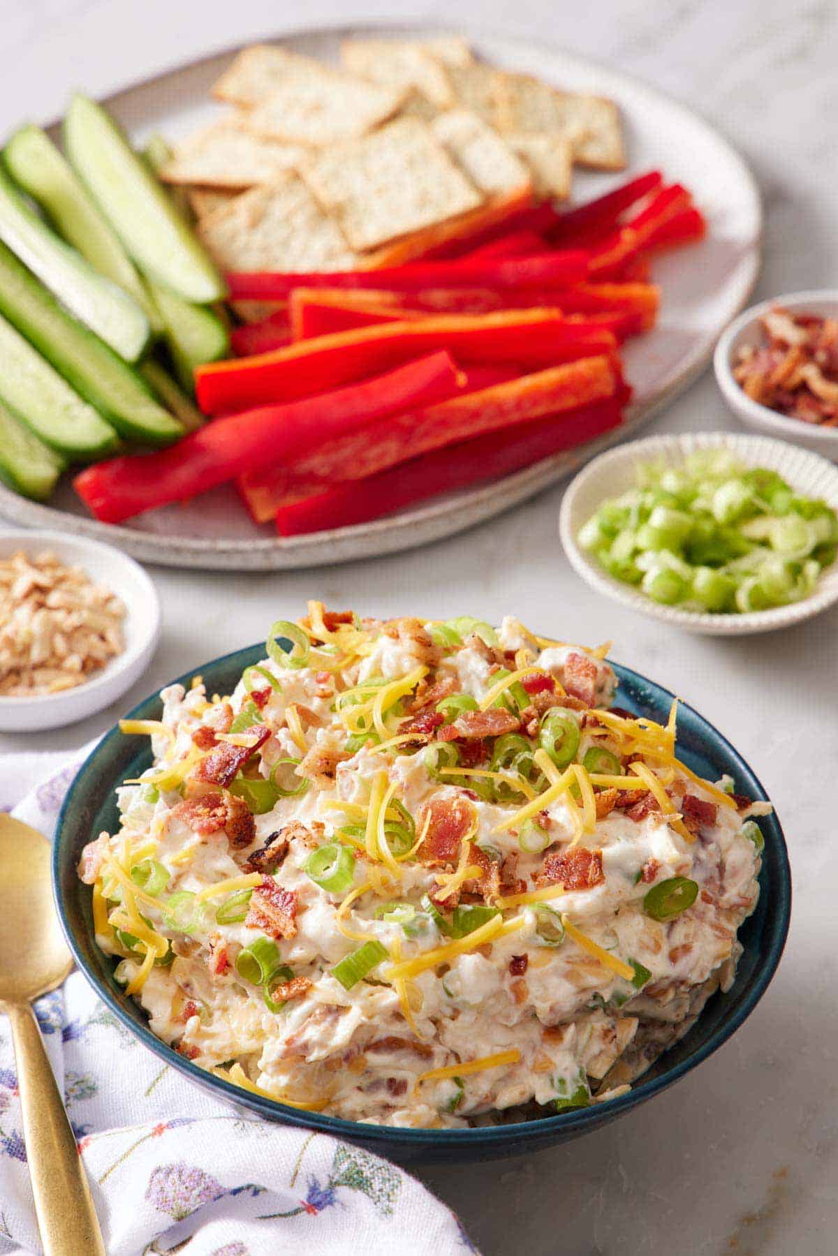 A bowl of million dollar dip with a platter in the background with cucumbers, peppers, and crackers. More ingredients in individual bowls off to the side.